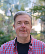 A man with short gray hair combed over smiles at the camera in front of green landscaping.