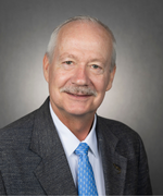 A man with thin white hair smiles at the camera wearing a suit jacket and a light blue tie.