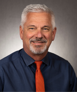 A man with gray hair smiles at the camera wearing a dark blue collared shirt and an orange tie.