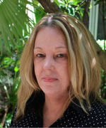 A woman with blond hair and a dark blouse smiles at the camera in the shade