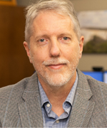 A man with gray hair smiles at the camera wearing a gray suit