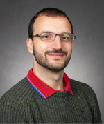 A man with short brown hair smiles at the camera wearing dark green sweater and red polo shirt.