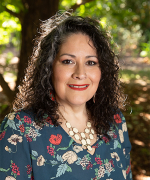 A woman with long black hair smiles at the camera wearing a flower-pattern blouse and white octagon necklace.