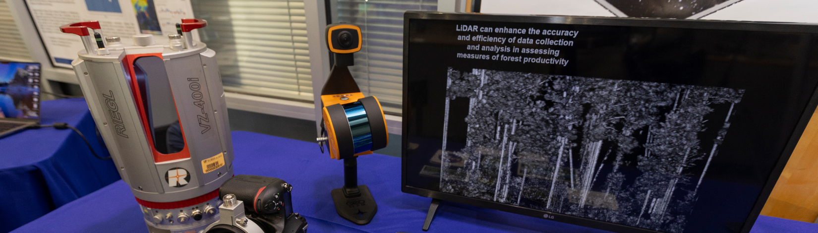 A piece of machinery that has a rotating camera attached sits on a table next to a computer screen displaying a 3 dimensional picture of a forest.